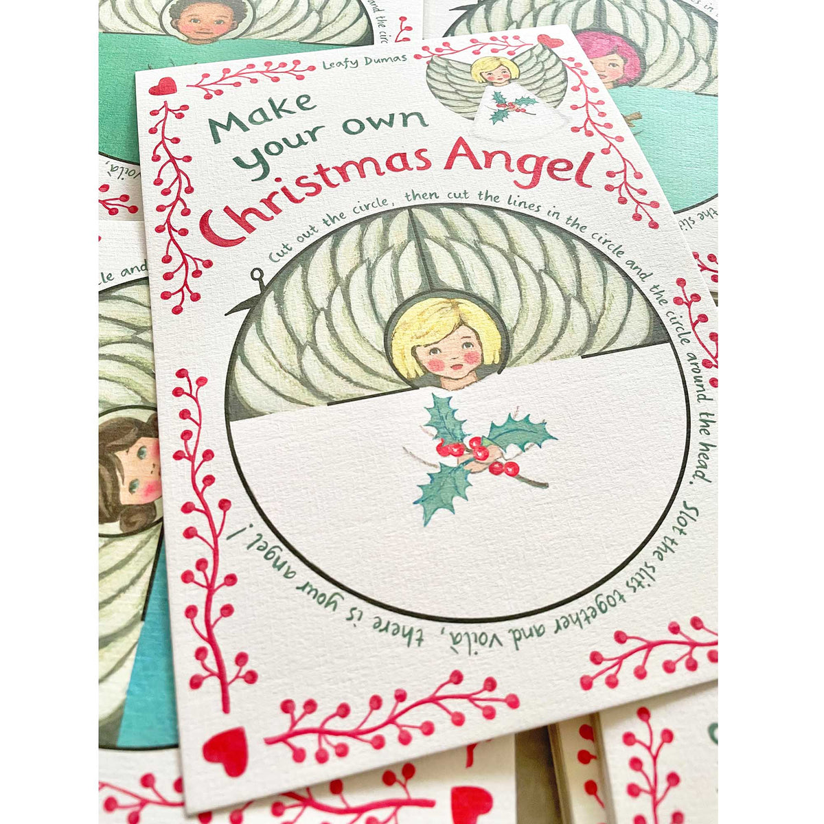A handmade Christmas card kit titled 'Make your own Christmas Angel' by Leafy Dumas, featuring a vintage-style illustration of an angel with gold hair and a green and white winged body, surrounded by red berries and holly on an off-white card.